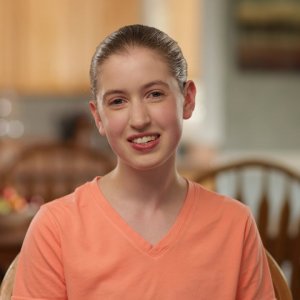 A young person with light skin and brown hair pulled back smiles while sitting indoors. They are wearing an orange V-neck shirt, with a blurred kitchen and dining area in the background.